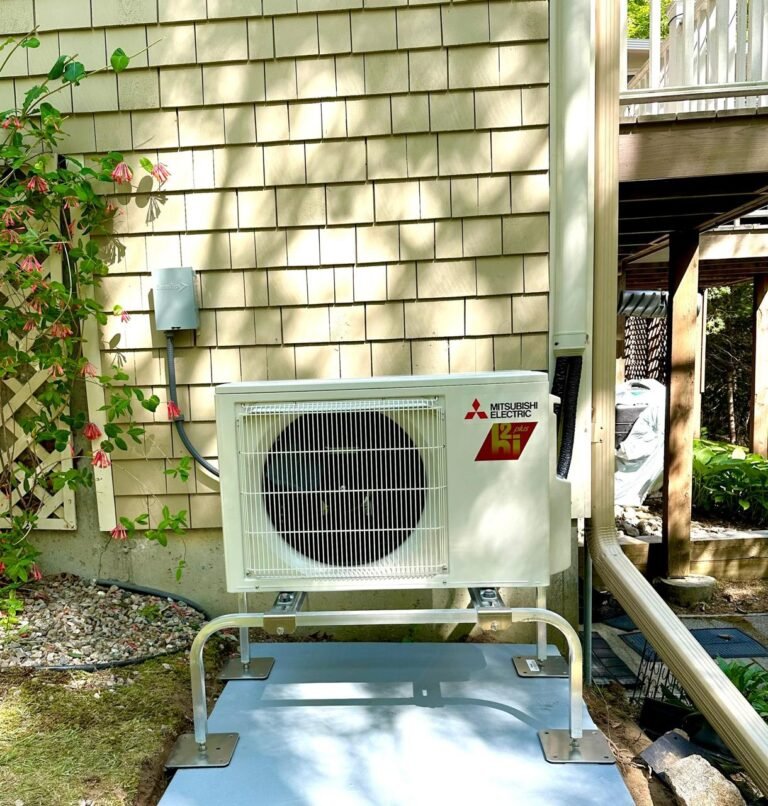 Mitsubishi Electric Hyper-Heat outdoor unit on a raised stand beside a Cape Cod shingle home